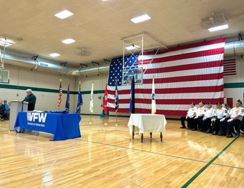 Speaker at a podium on the left, veterans on the left, and a giant American flag in the back